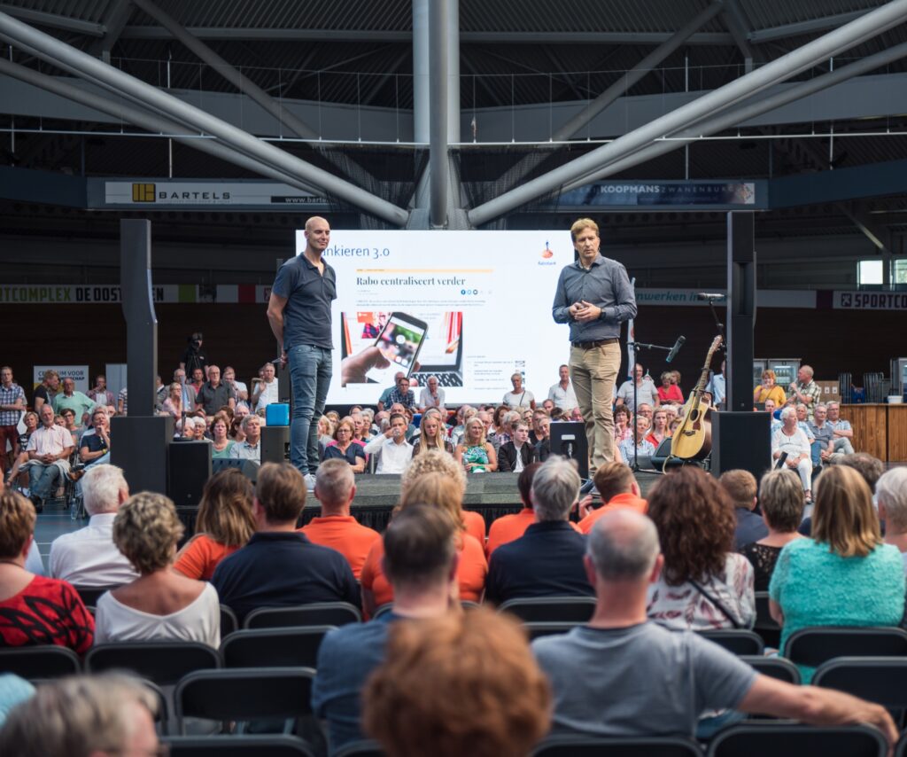 Op een podium staat een presentator en Maarten van der Weijden tijdens een zakelijk evenement